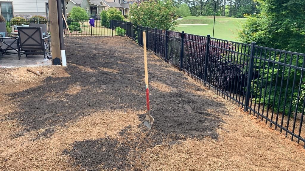 Yard covered in brown mulch with a shovel, surrounded by a black fence and greenery.