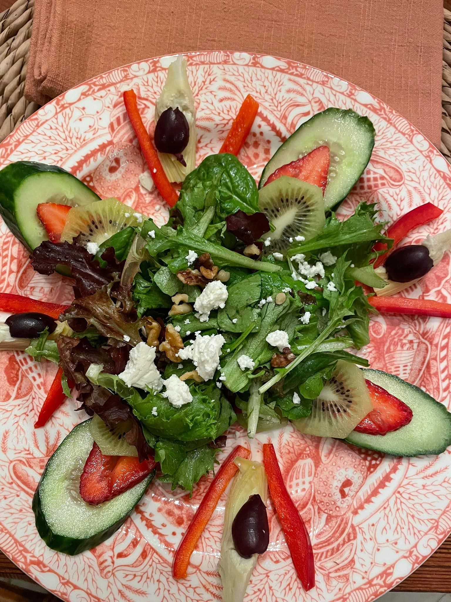 Decorative salad with greens, kiwi, strawberries, black olives, carrots, and crumbled cheese on a patterned plate