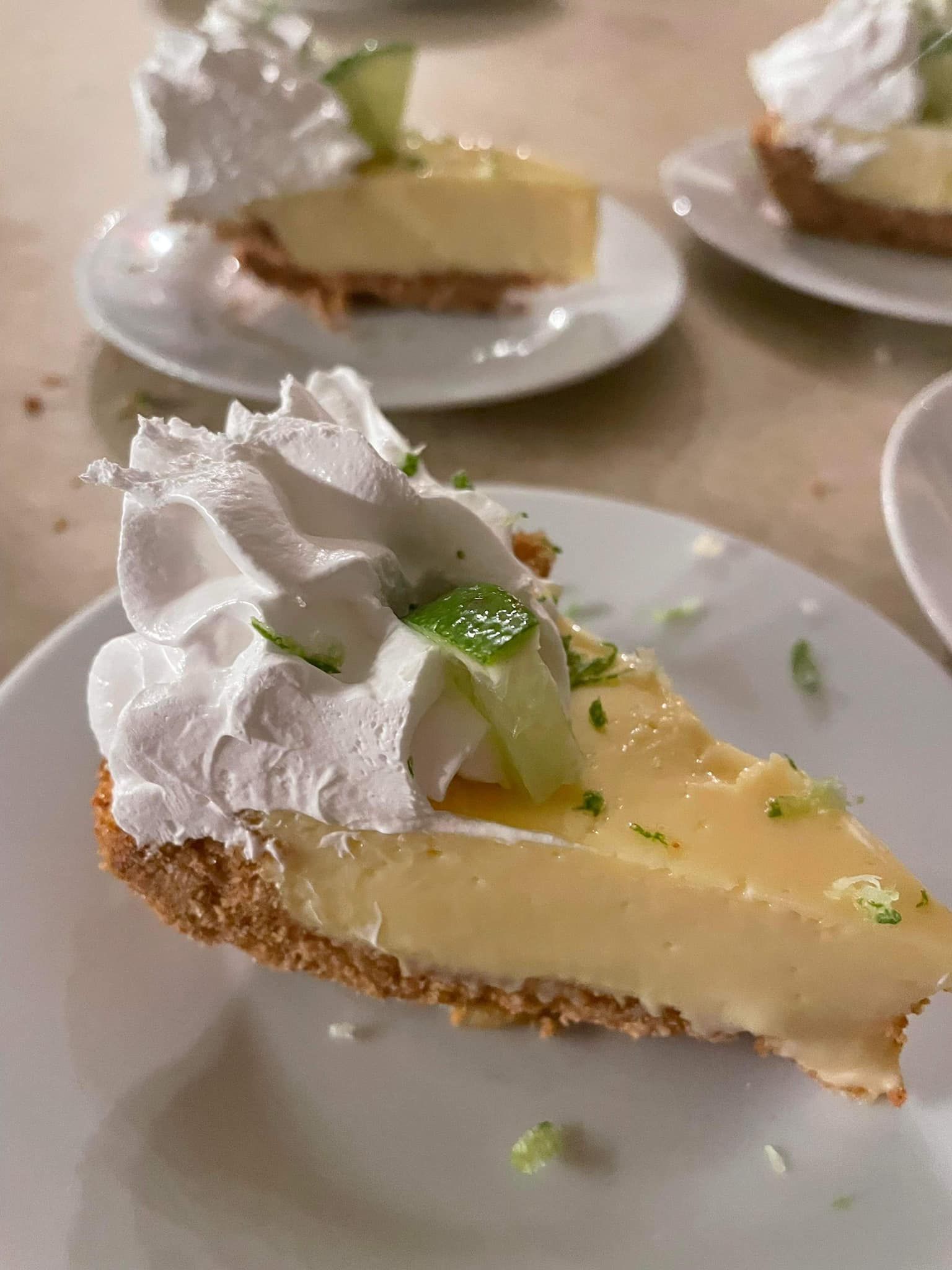 Slice of key lime pie with whipped cream and lime garnish on a white plate, with more slices blurred behind