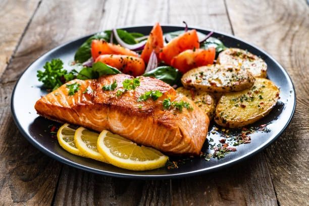 Grilled salmon with lemon slices, roasted potatoes, and salad on a black plate on a wooden table