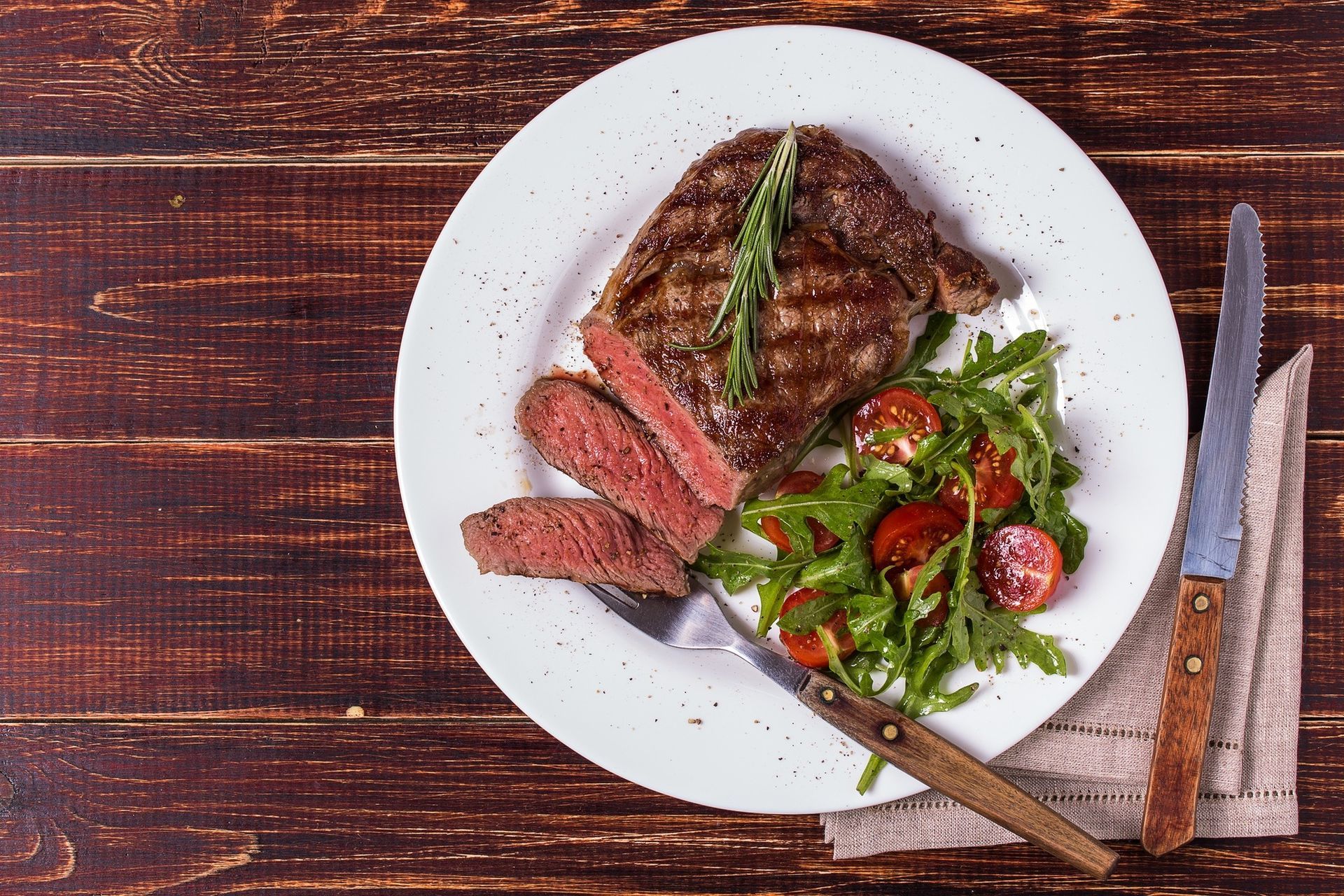 Steak dinner with sliced meat, salad, and asparagus on a white plate with knife and fork on a wooden table