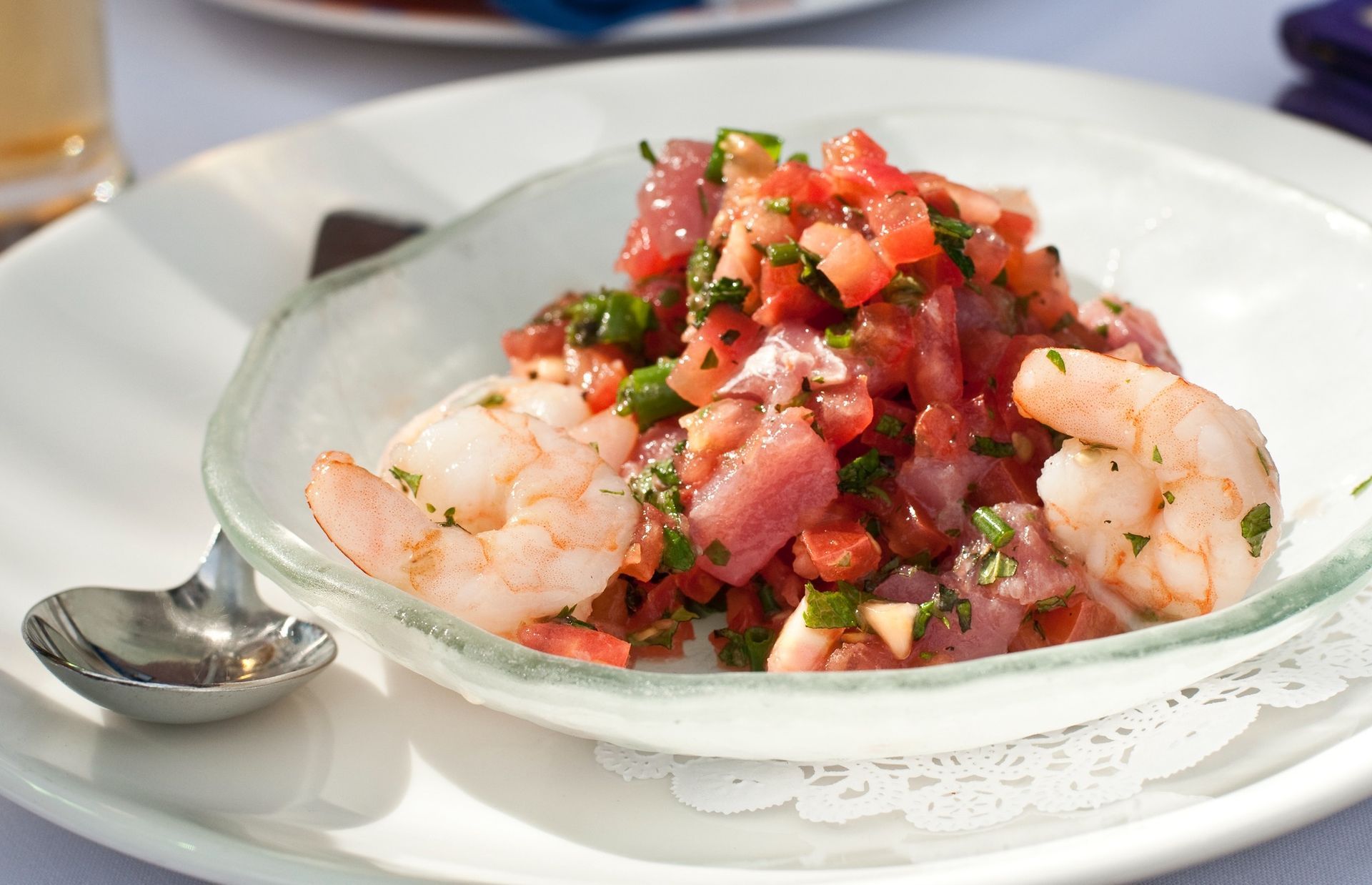 Shrimp ceviche in a glass bowl topped with diced tomatoes, served on a white plate with a spoon