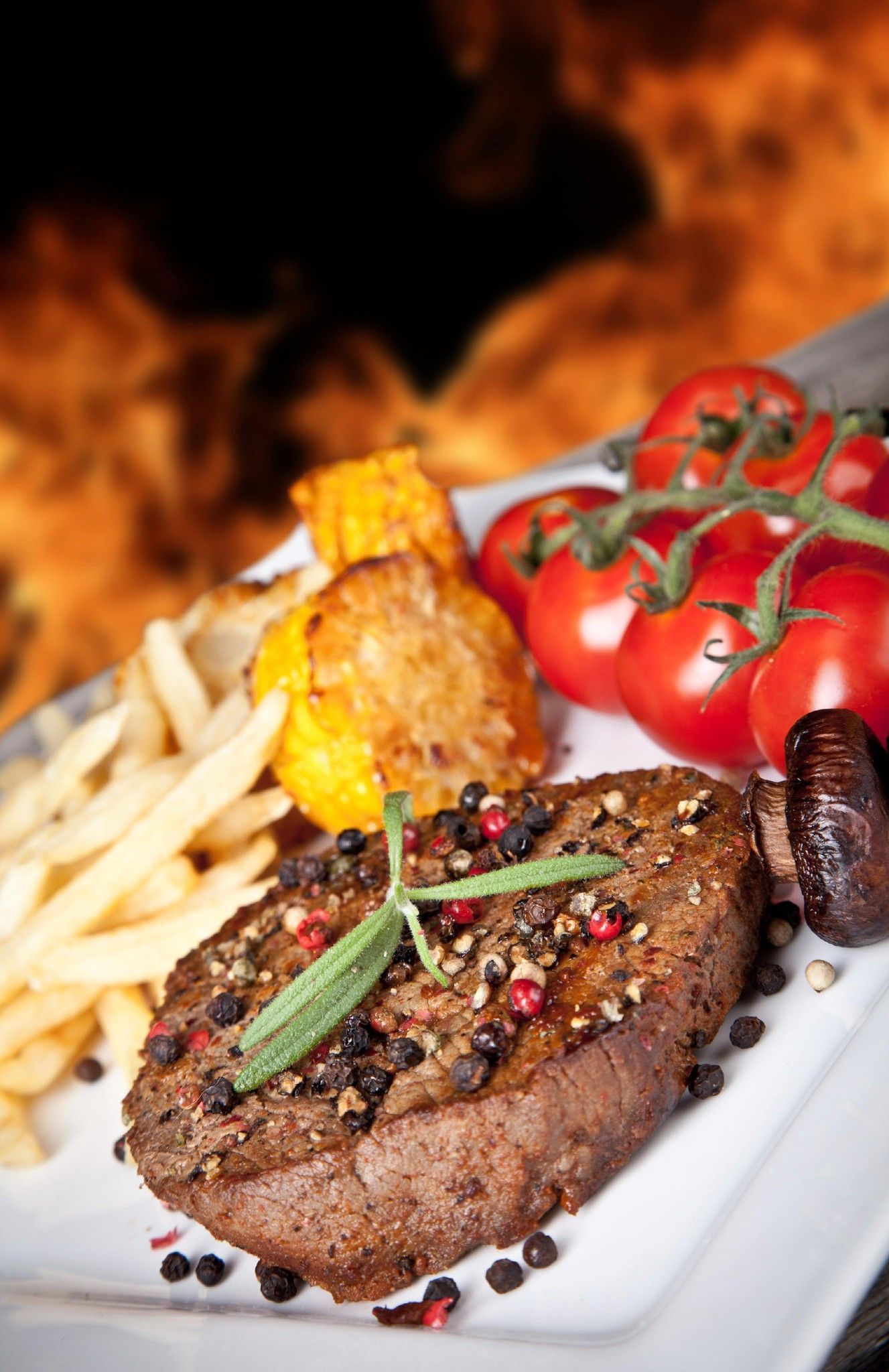 Grilled steak with fries, tomatoes, and sauce on a white plate with flames in the background
