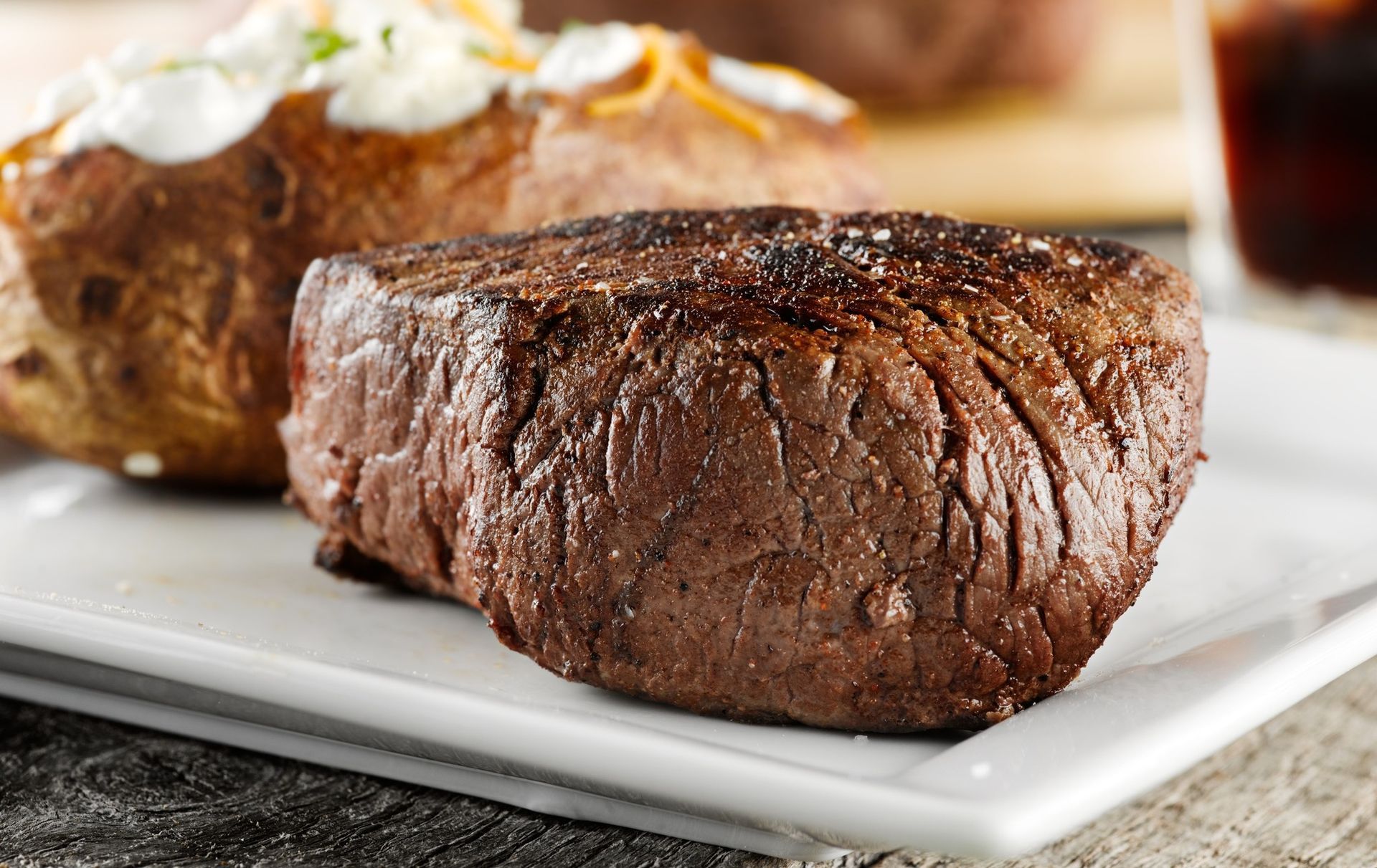 Grilled steak on a white plate with a baked potato and a glass of red wine in the background