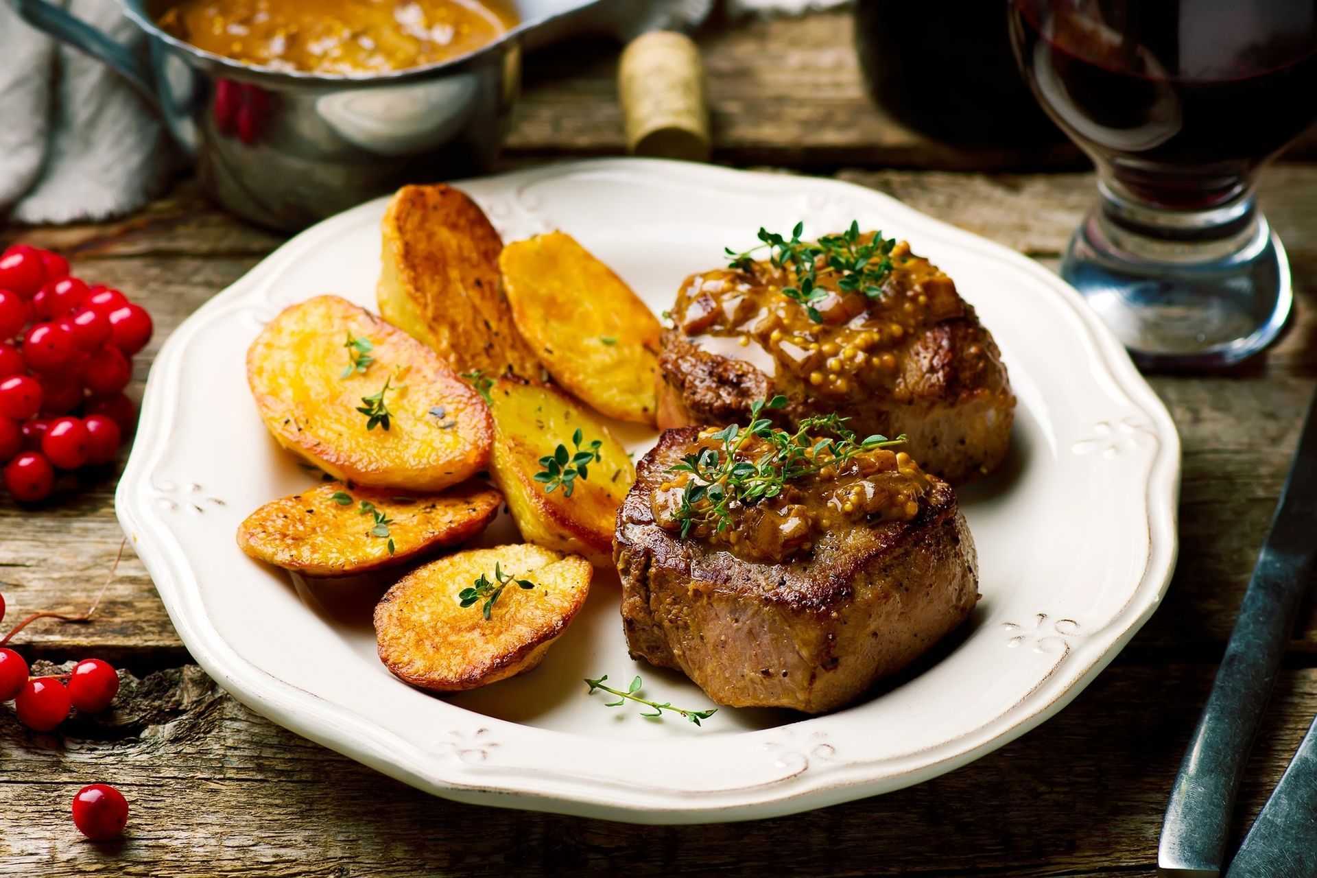 Grilled steak with roasted potatoes and cherries on a white plate, with sauce and a glass of red wine nearby