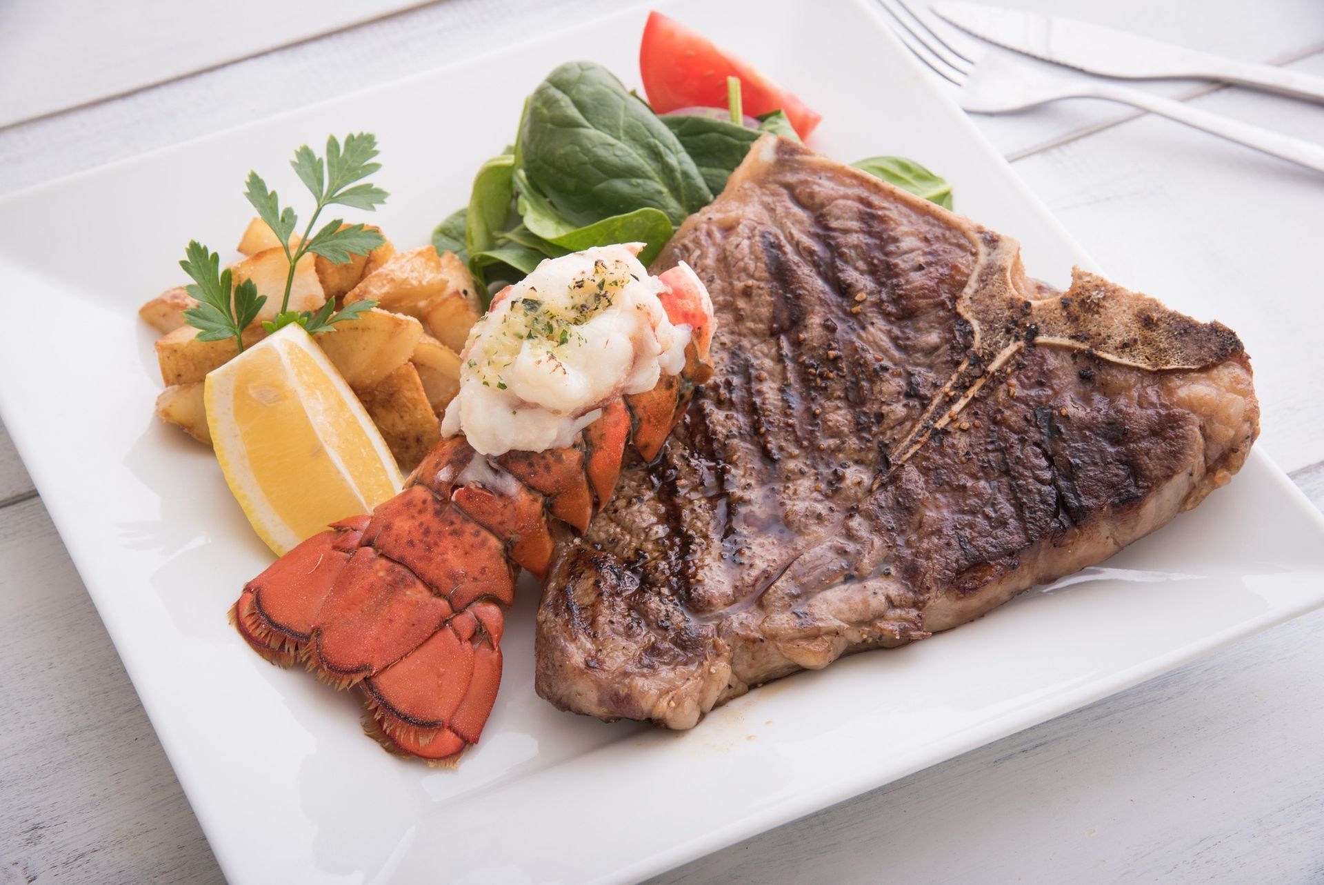 Grilled steak with lobster tail, lemon wedge, and vegetables on a white plate