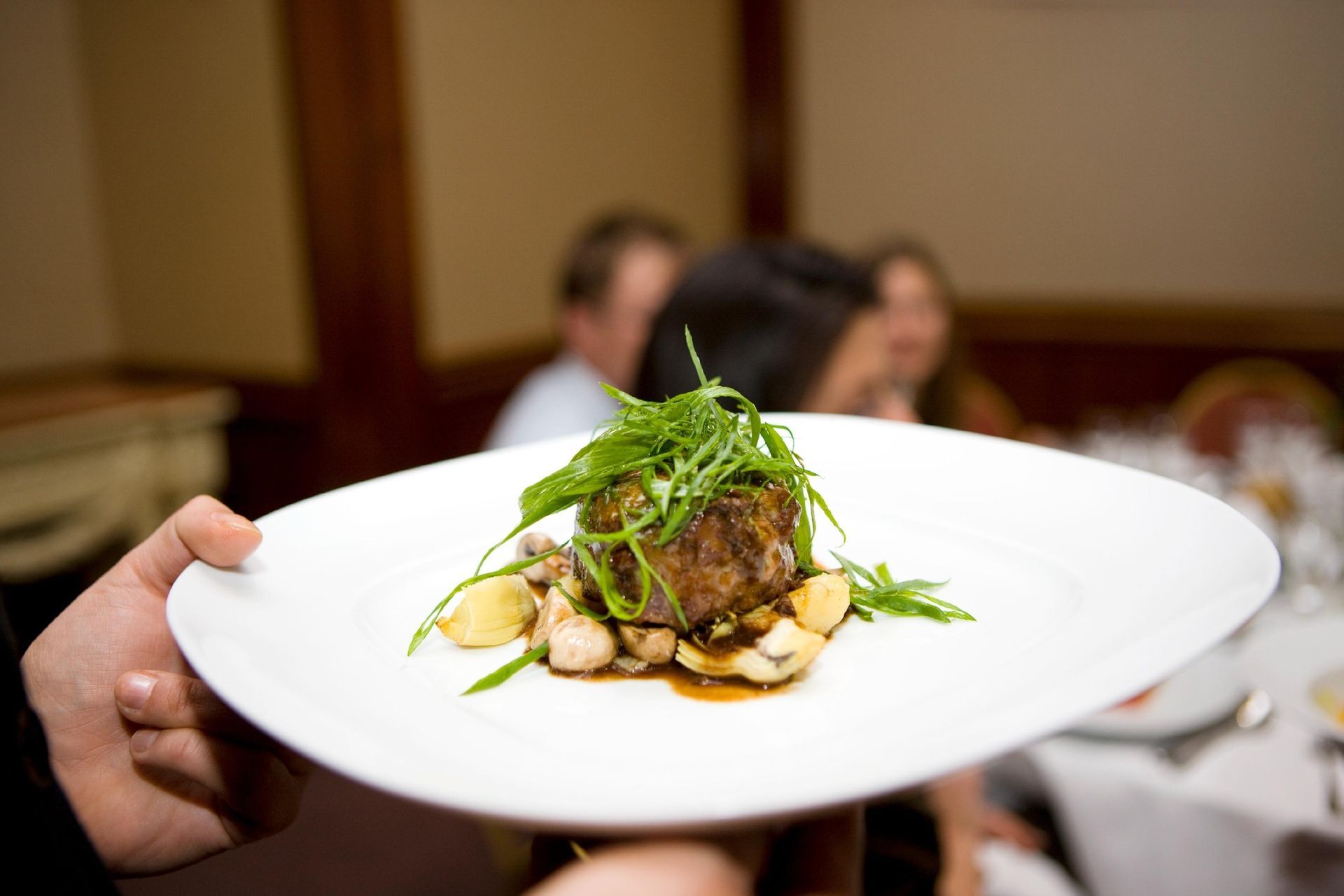Plated gourmet dish held at a dining table, topped with greens and sauce, with blurred people in background