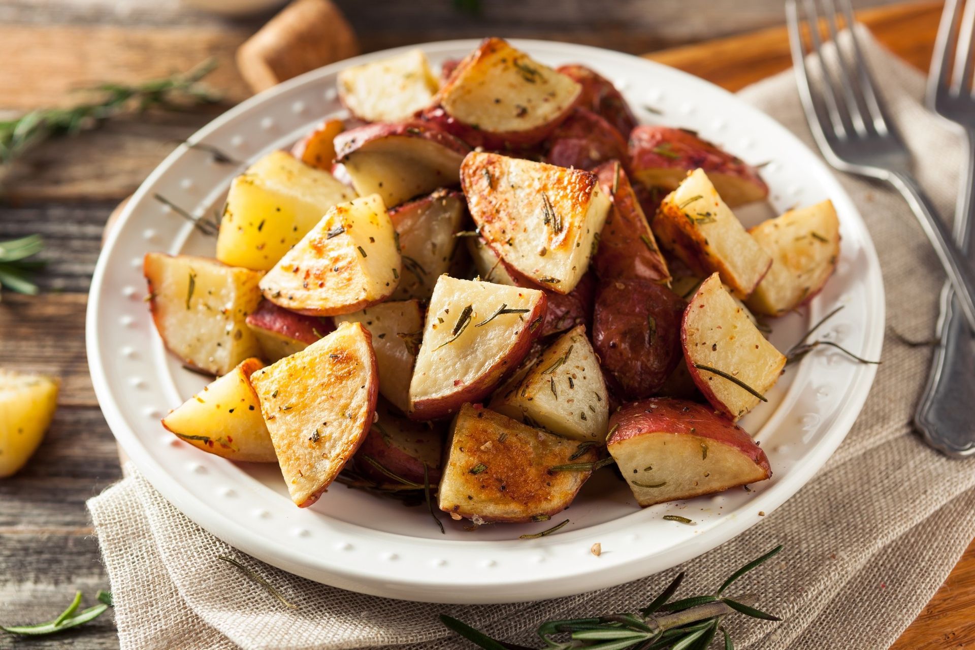 Roasted potato wedges on a white plate with herbs and seasoning, served with a fork on a linen napkin.
