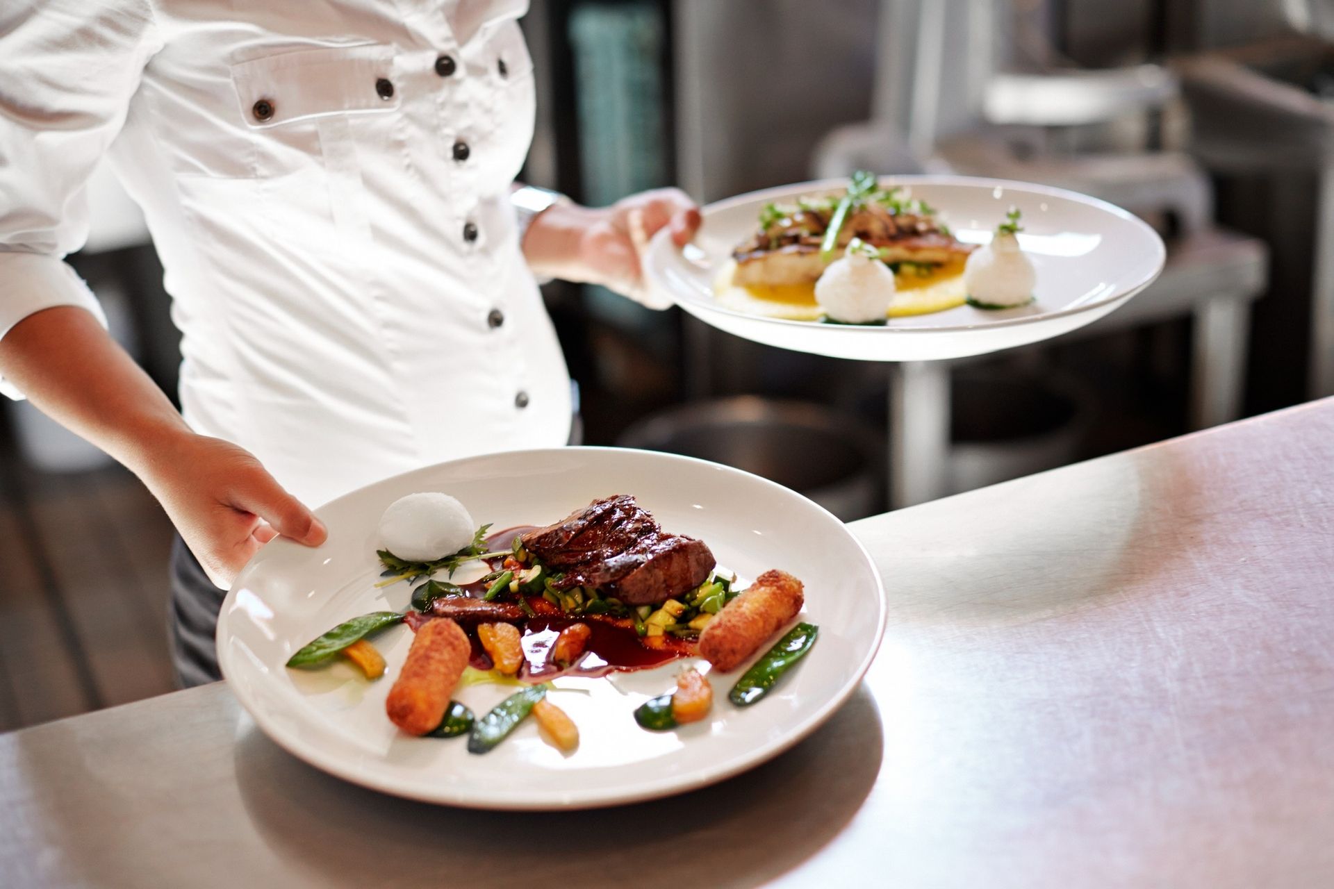 Chef carrying two plated gourmet dishes in a restaurant kitchen