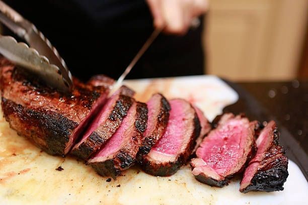 Sliced medium-rare steak on a cutting board, with tongs holding the roast and a blurred kitchen background