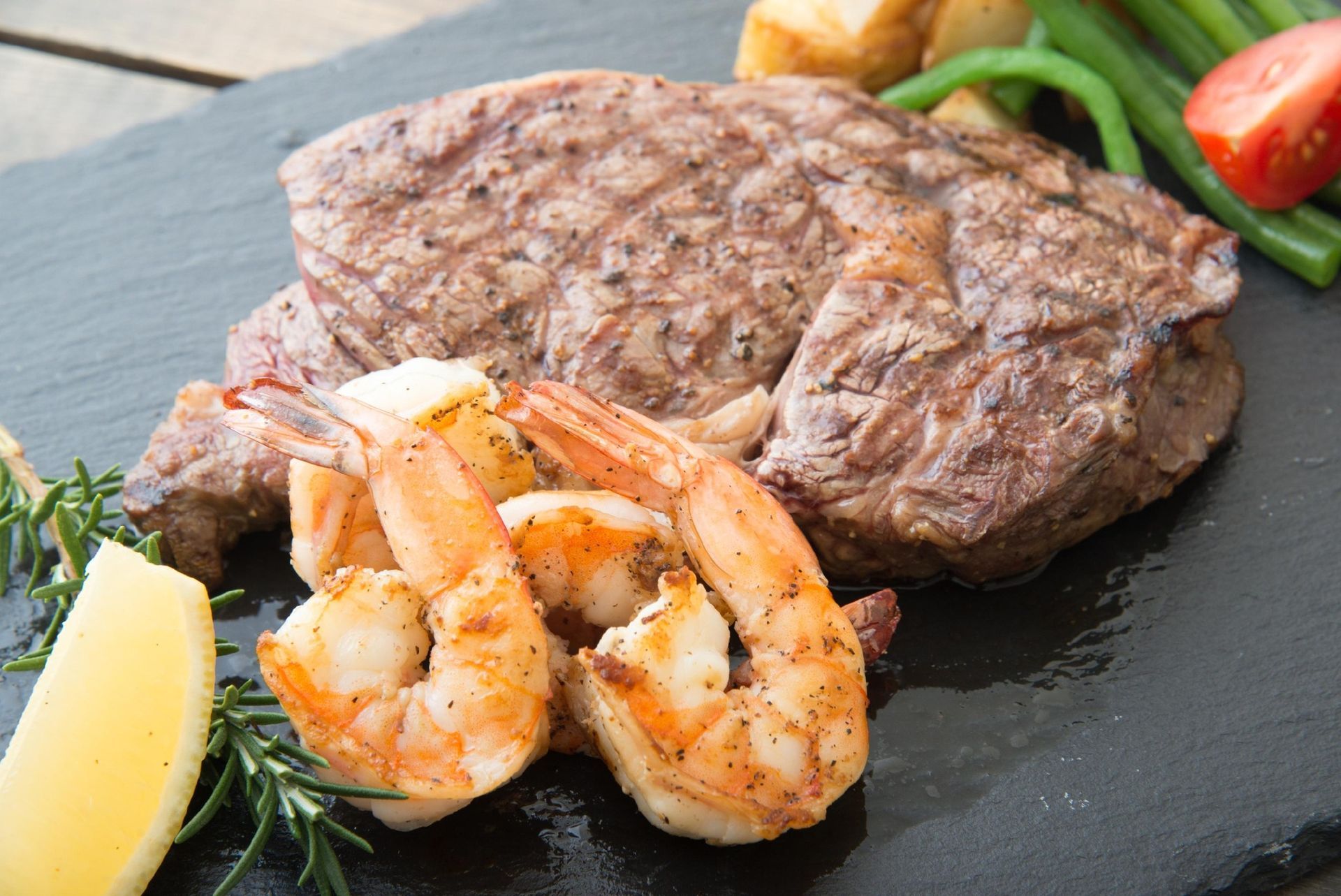 Grilled steak with shrimp, lemon wedge, and vegetables on a black slate plate