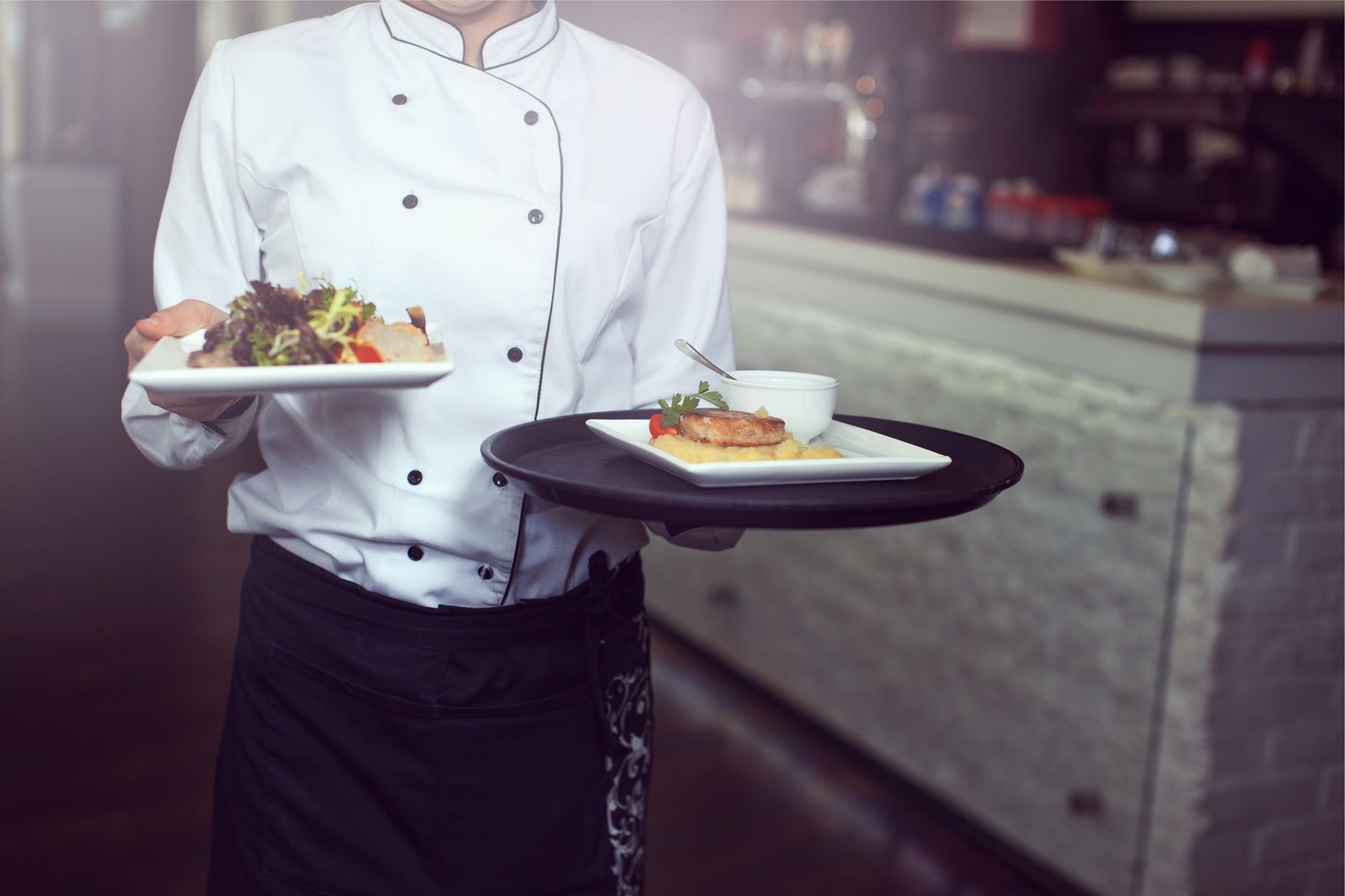 Waiter carrying plates of food in a restaurant, wearing a white uniform and black apron