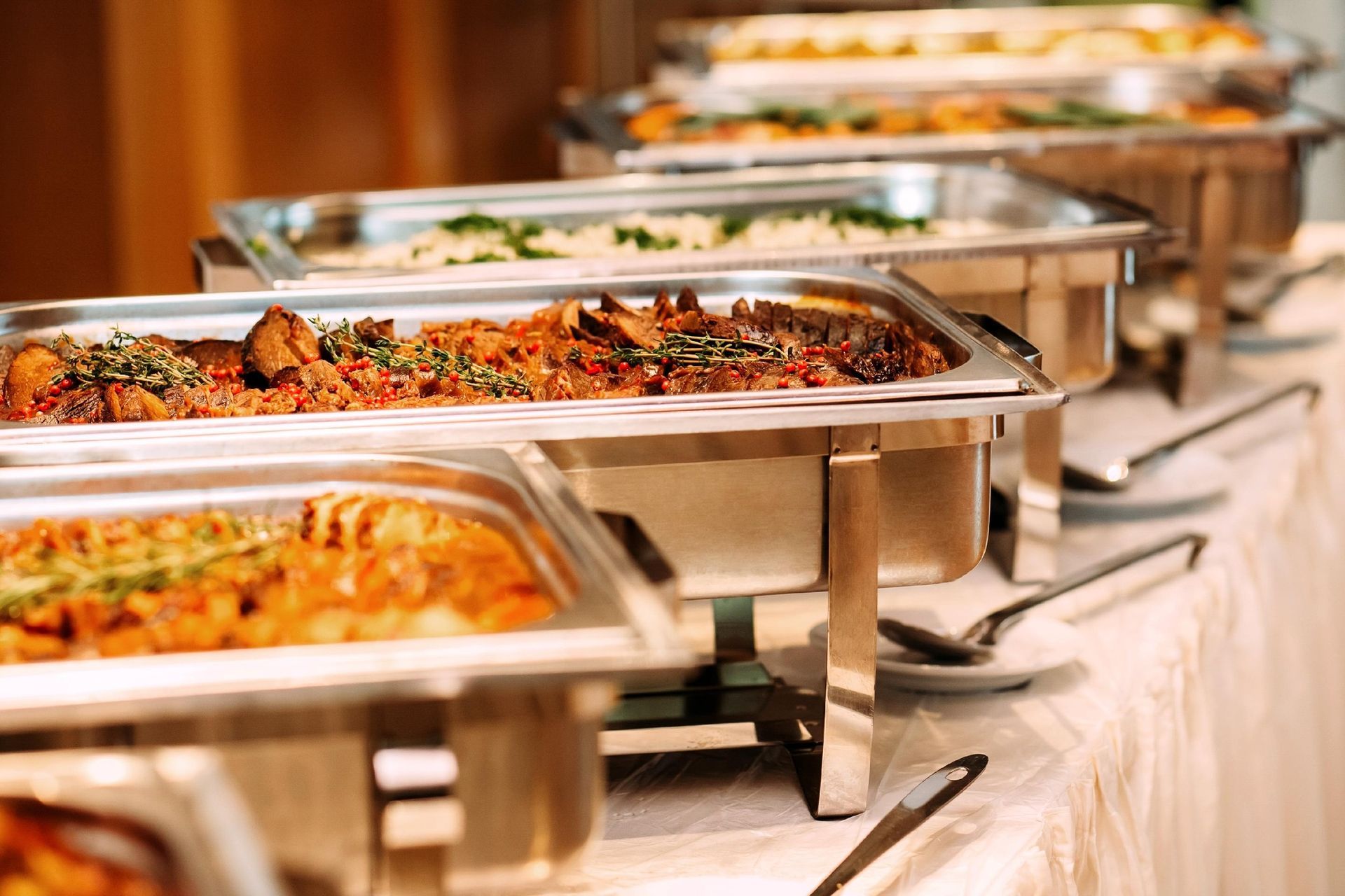 Buffet table with several stainless-steel trays of assorted hot dishes under warming lights