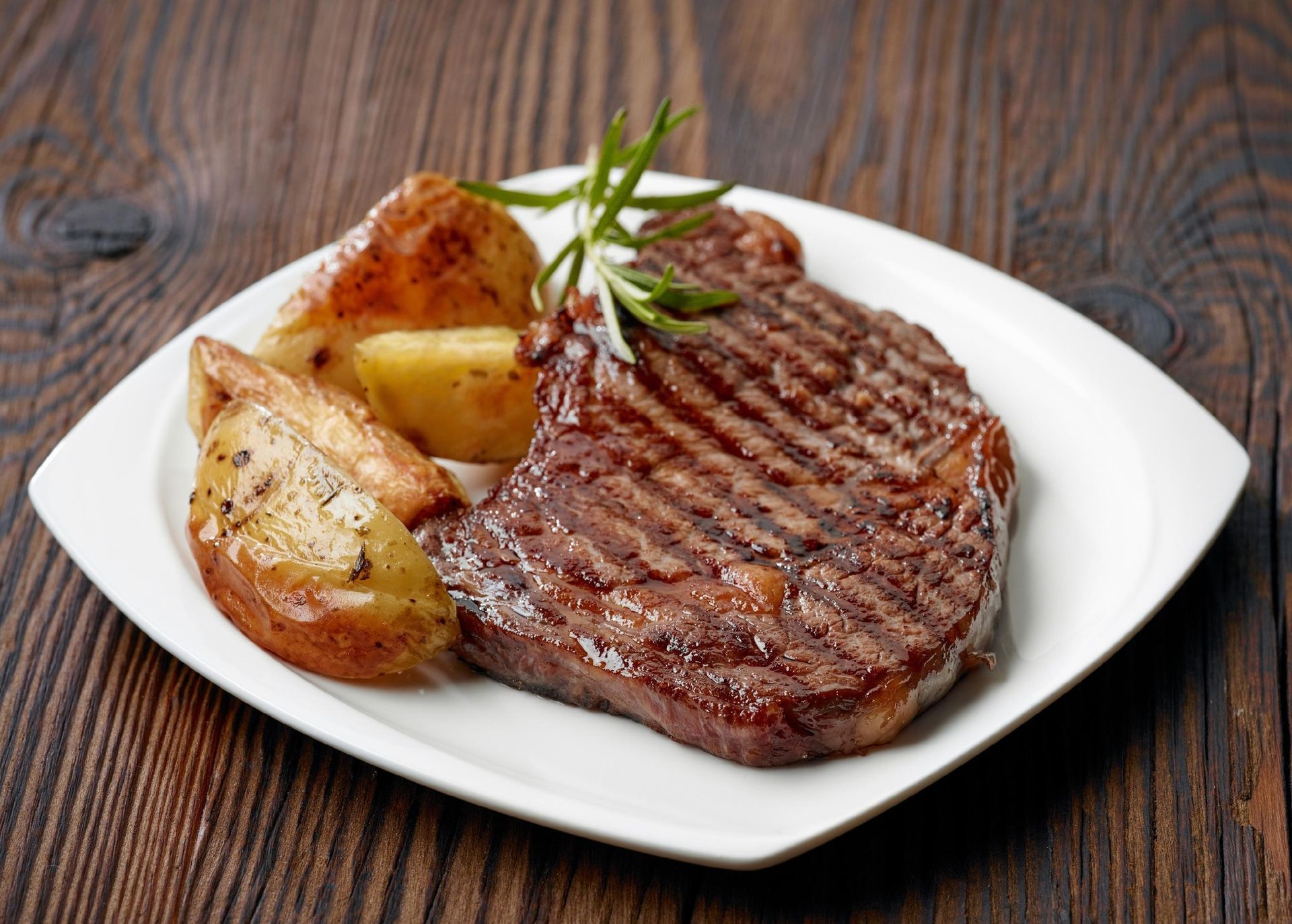 Grilled steak with roasted potato wedges on a white plate, garnished with rosemary, on a wooden table