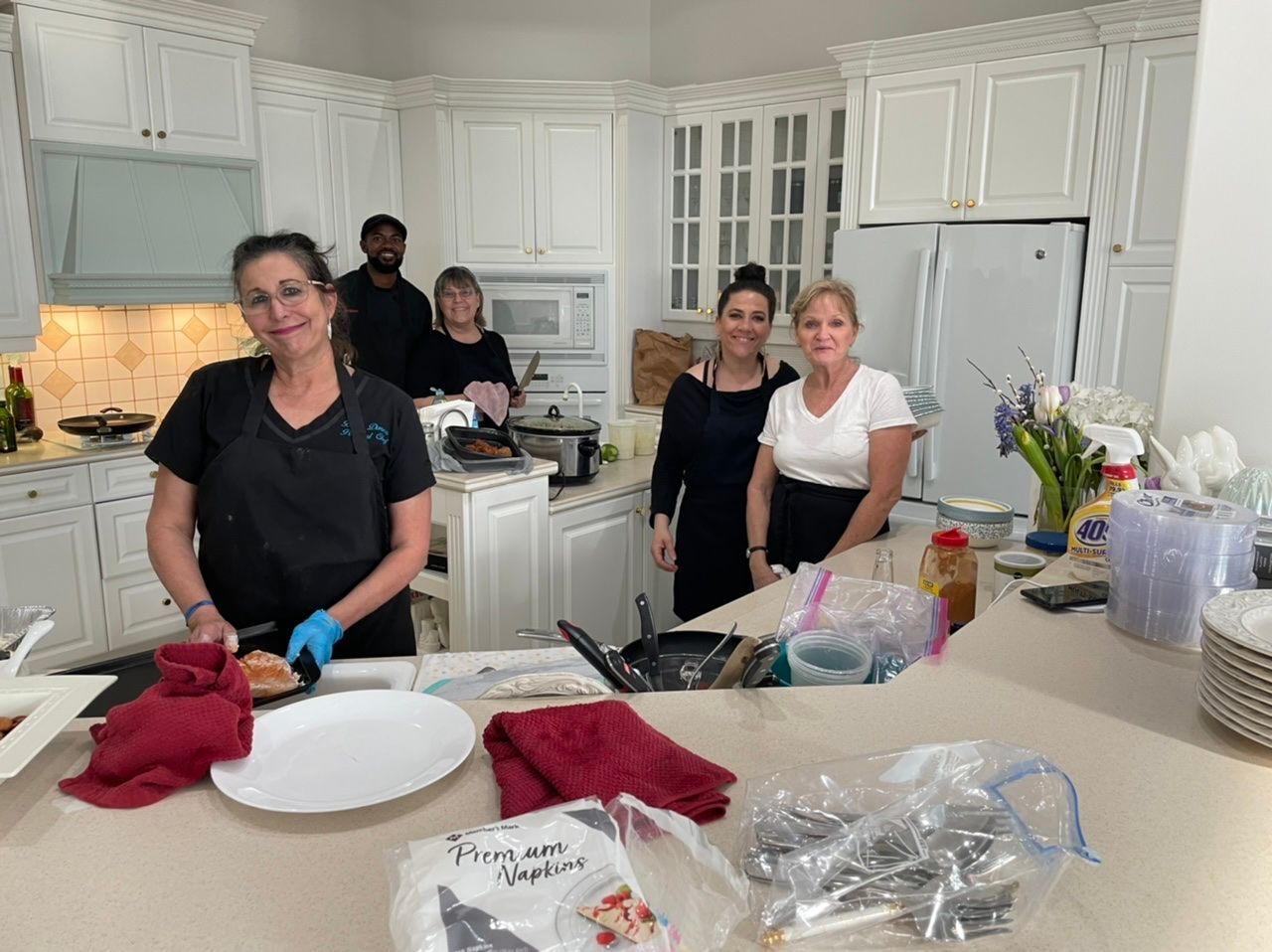 People in a bright white kitchen preparing food at a counter with plates, gloves, and ingredients