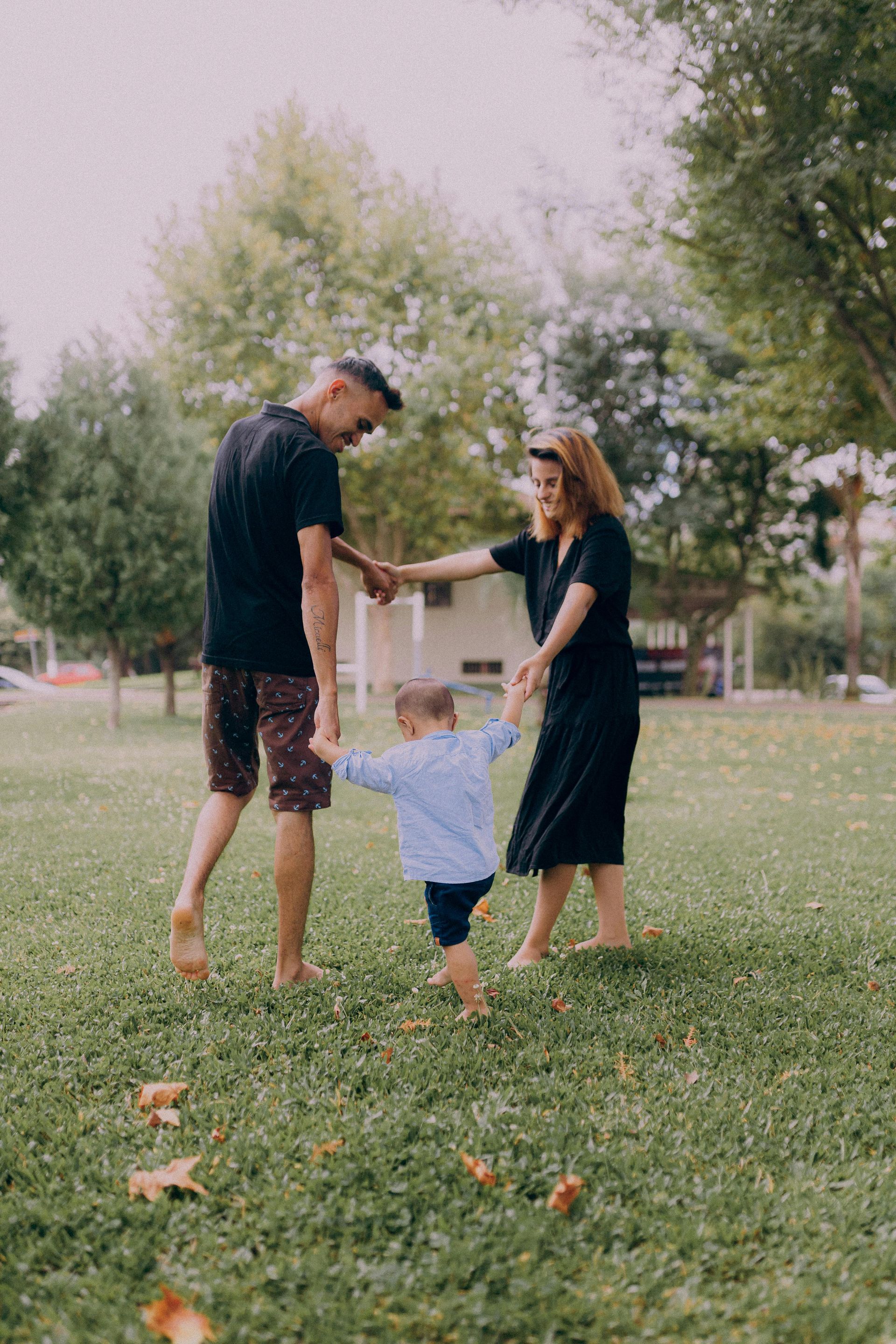 Three people holding hands in a grassy park, with trees and a house in the background.