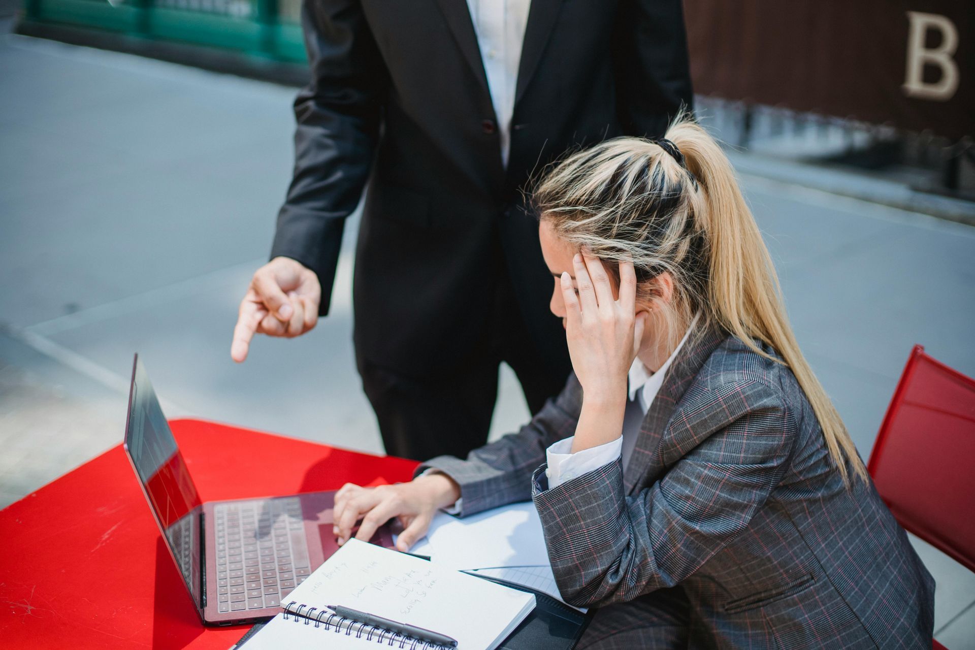 Woman using a laptop at a red outdoor table while a man stands nearby pointing at the screen