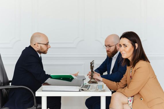 Three people in a meeting across a white table, discussing documents and a laptop in a bright office.