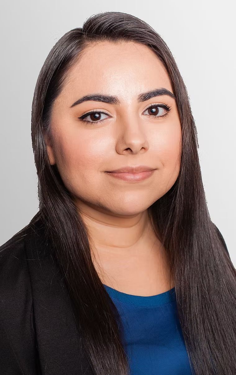 Studio headshot of a person with long dark hair wearing a black blazer and blue top, against a light gray background