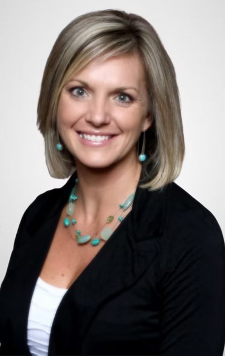 Smiling person in a black blazer, wearing turquoise jewelry, against a light gray background