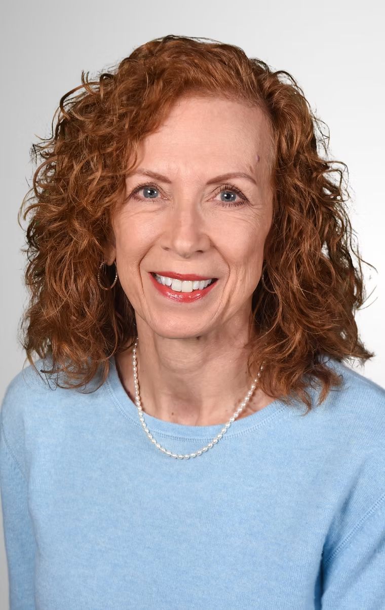 Smiling woman with curly red hair wearing a light blue sweater and pearl necklace on a white background