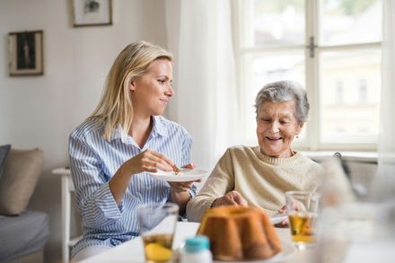Two women share tea at a table in a bright kitchen, one serving the other, both smiling.