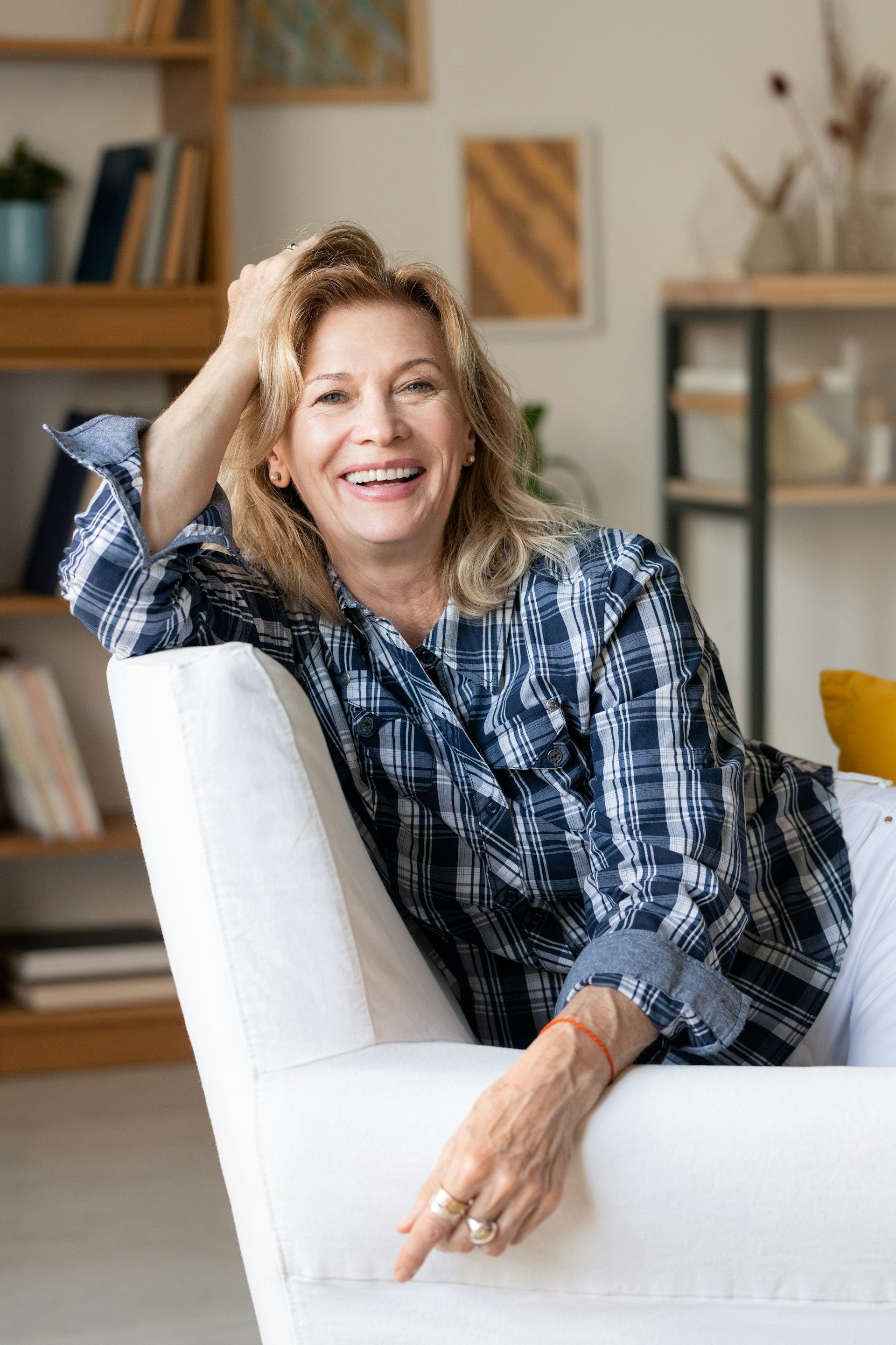 Smiling woman in a blue plaid shirt sitting on a white chair in a cozy living room