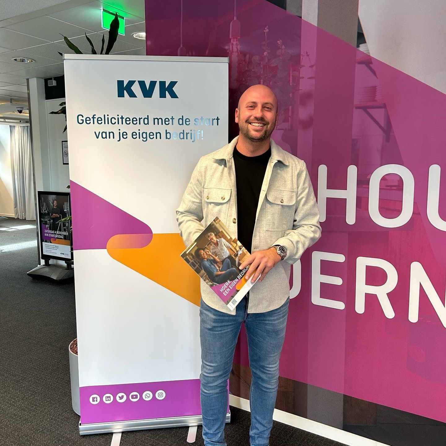 Een glimlachende man met een brochure in zijn hand staat naast een spandoek van KVK.