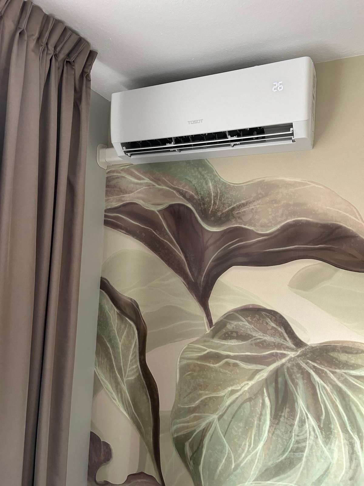 Witte airconditioner op een muur met een patroon, met links een bruin gordijn.