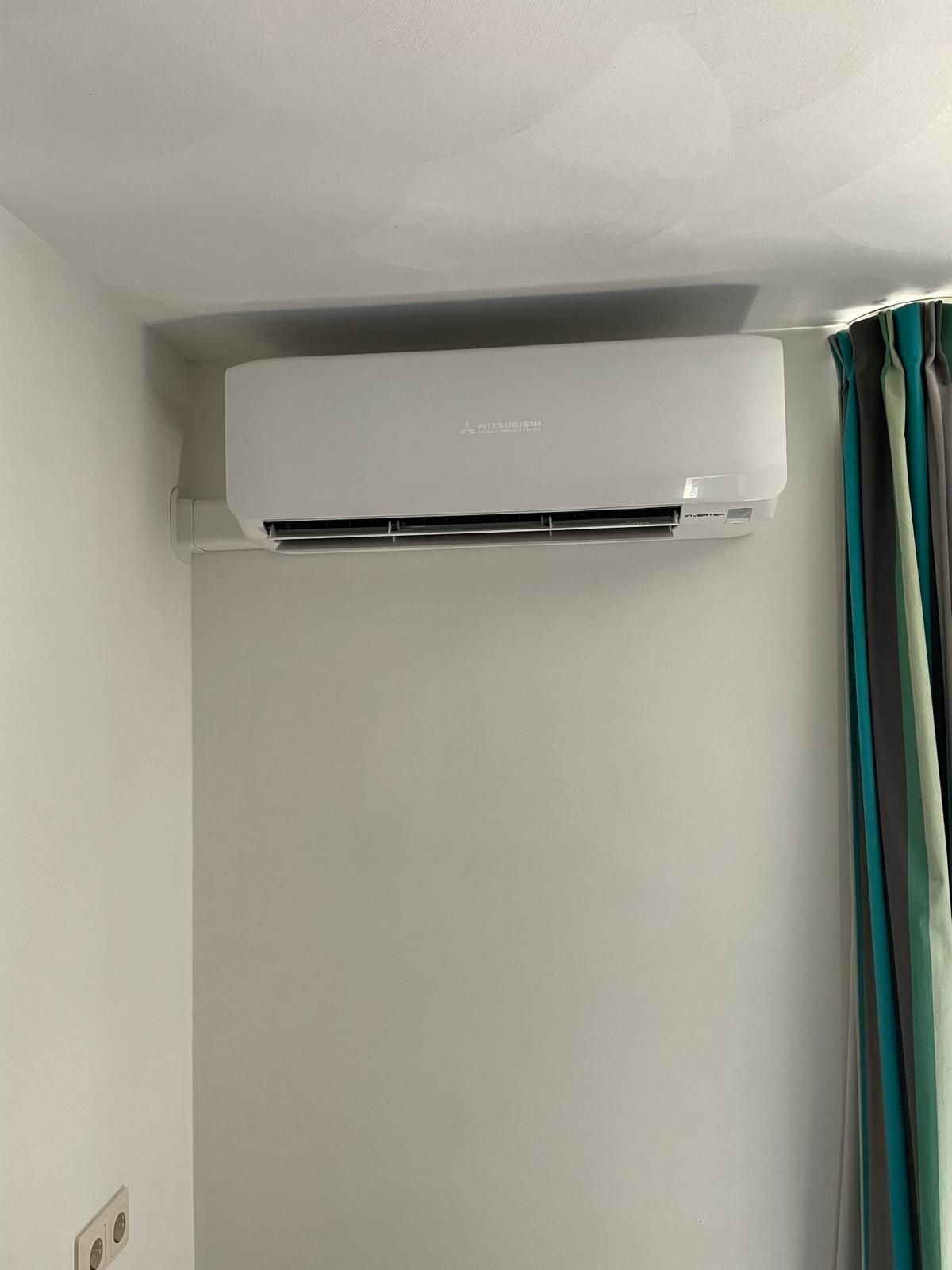 Witte, aan de muur gemonteerde airconditioner boven een lichtgrijze muur, met turquoise gordijnen rechts ervan.