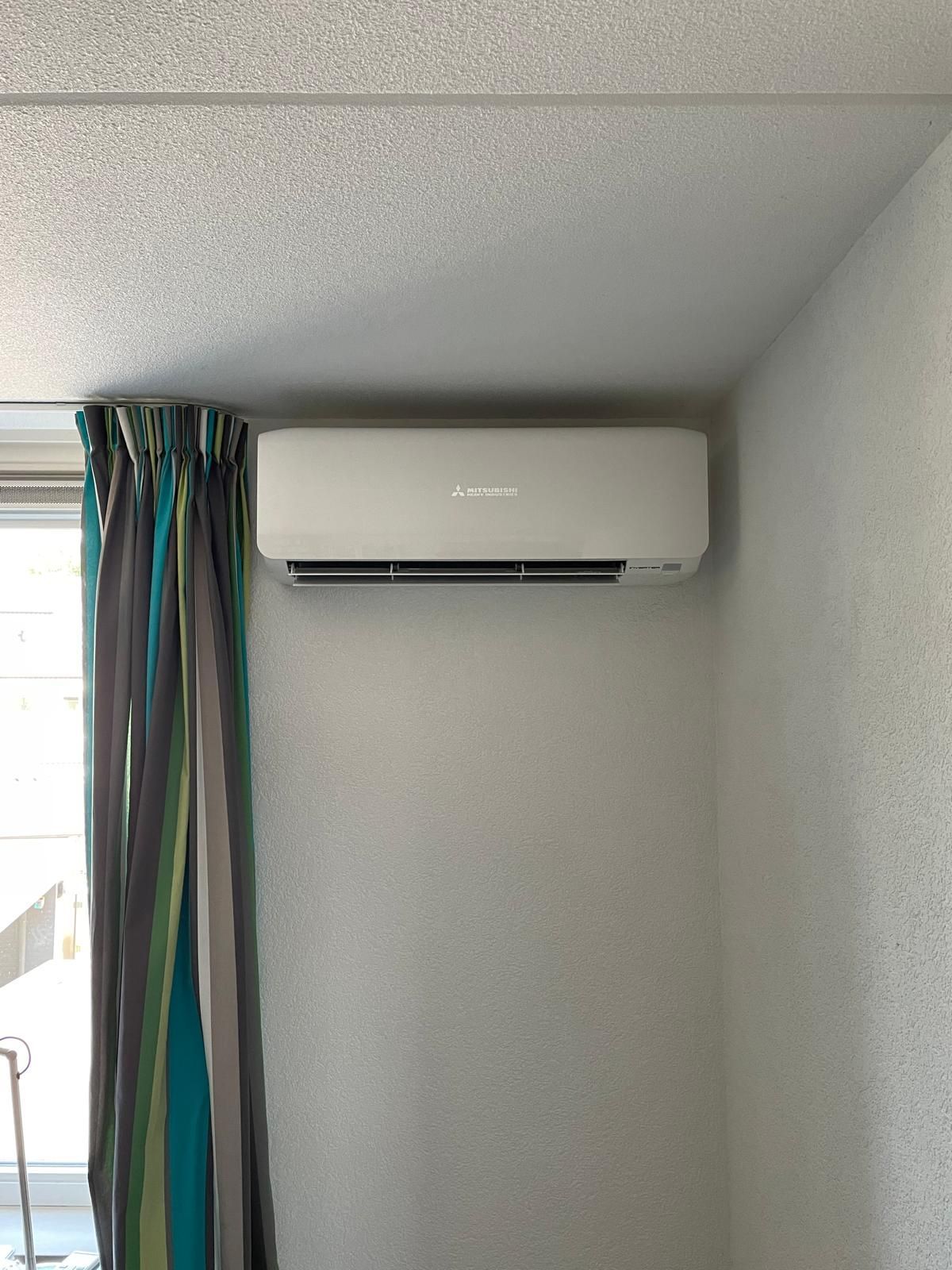 Witte airconditioningunit gemonteerd aan een lichtgrijze muur naast een raam met blauw-groen gestreepte gordijnen.