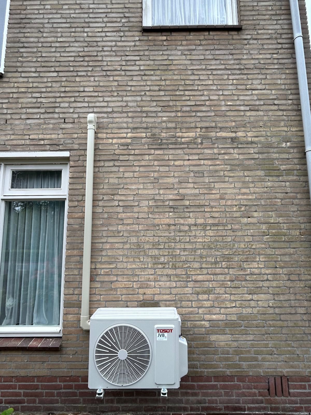 Airconditioningunit gemonteerd op een bakstenen muur met raam en regenpijp.