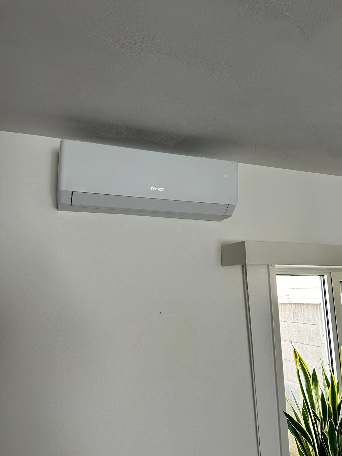Witte, aan de muur gemonteerde airconditioningunit boven een raam met uitzicht op groen.