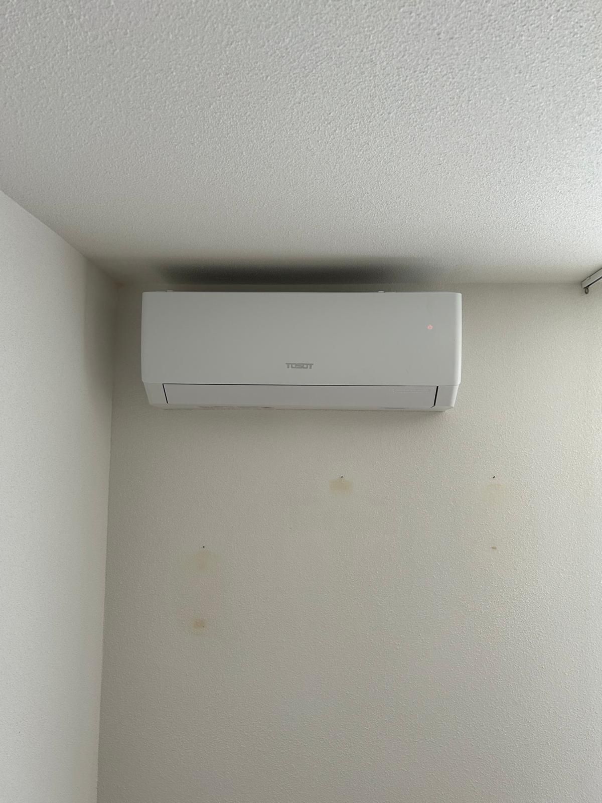 Witte airconditioningunit gemonteerd aan een witte, gestructureerde muur in een hoek.