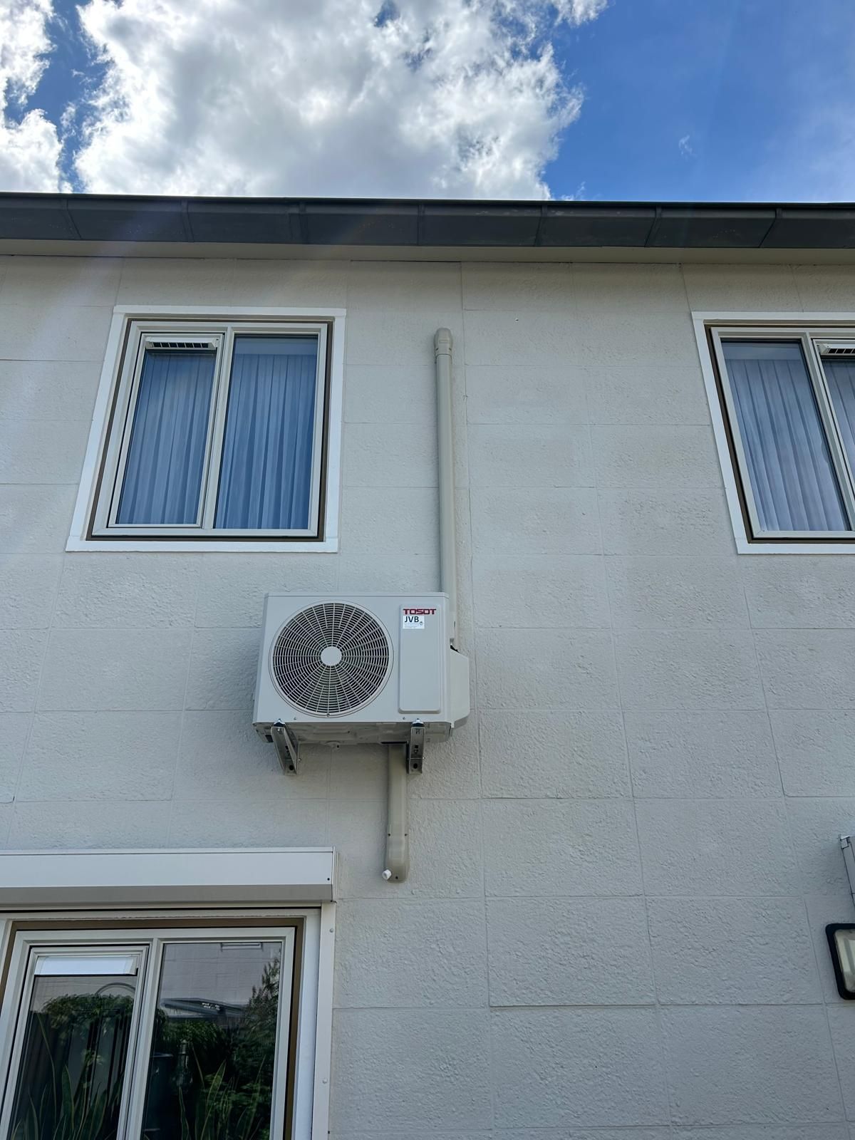 Airconditioningunit gemonteerd op een lichtgrijs gebouw met drie ramen. Een leiding loopt langs de buitenmuur omhoog.