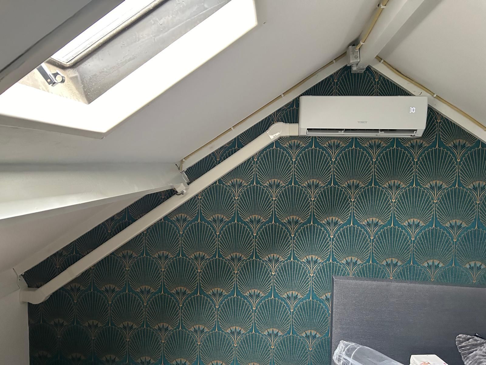 Airconditioning gemonteerd op een behang met een turquoise patroon, onder een schuin plafond met een dakraam.