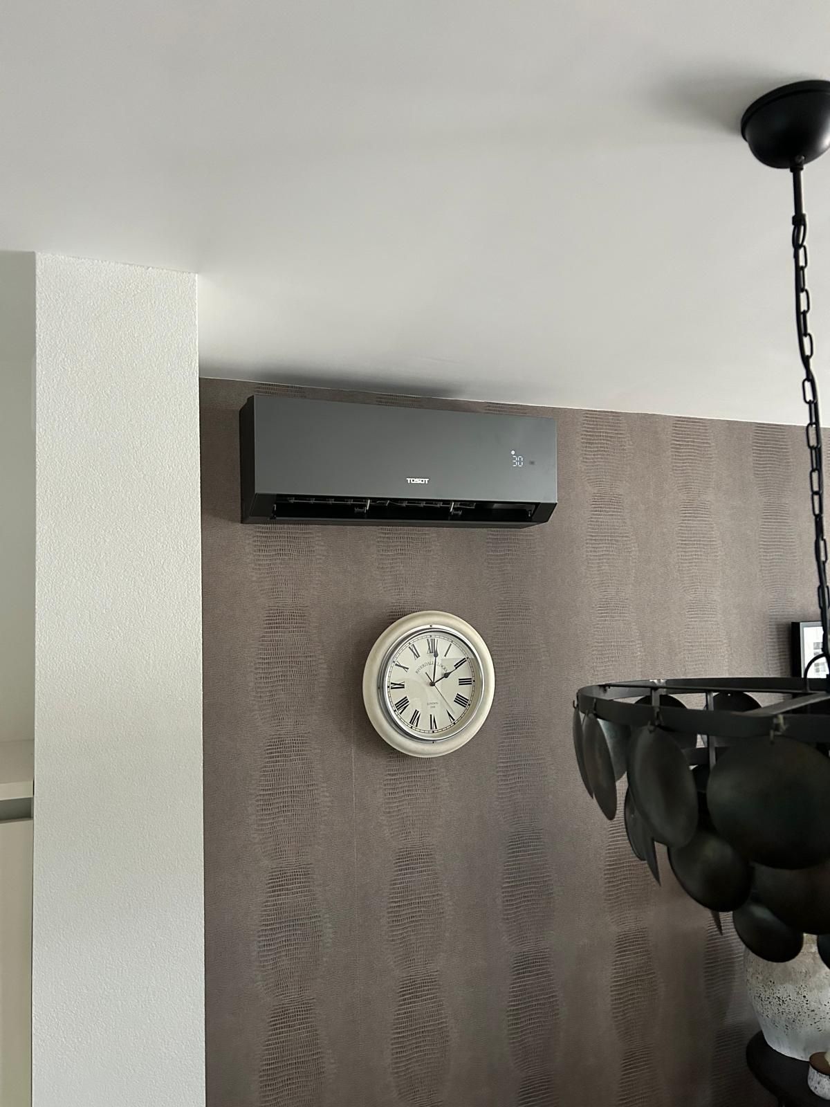 Een grijze airconditioner hangt boven een klok aan een bruine muur met structuurverf, en vlakbij hangt een kroonluchter.