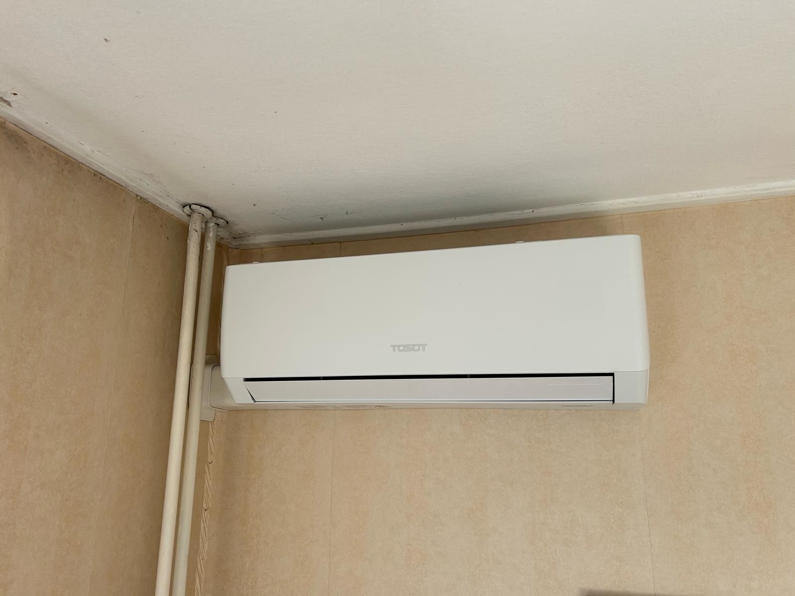 Witte airconditioningunit gemonteerd aan een beige muur nabij de hoek van het plafond, met zichtbare leidingen.
