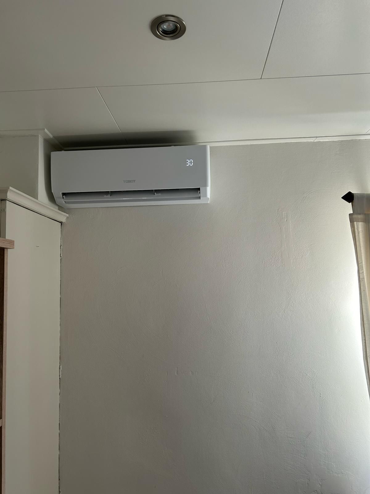 Witte airconditioner gemonteerd aan een lichtgrijze, gestructureerde muur in een kamer.
