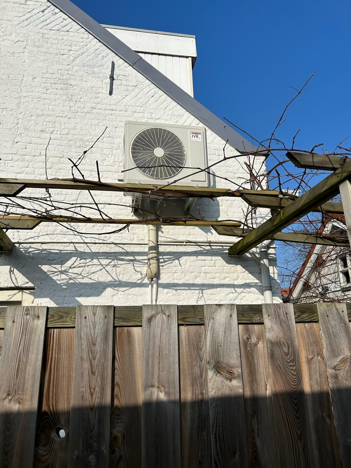 Wit gebouw met airconditioning, houten schutting en pergola onder een blauwe hemel.