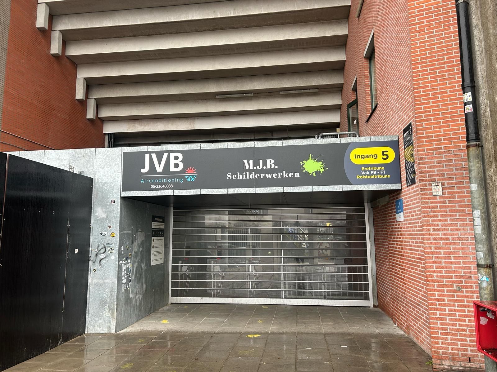 Ingang van JVB Schilderwerken onder de stadiontribune met een afgesloten beveiligingspoort.