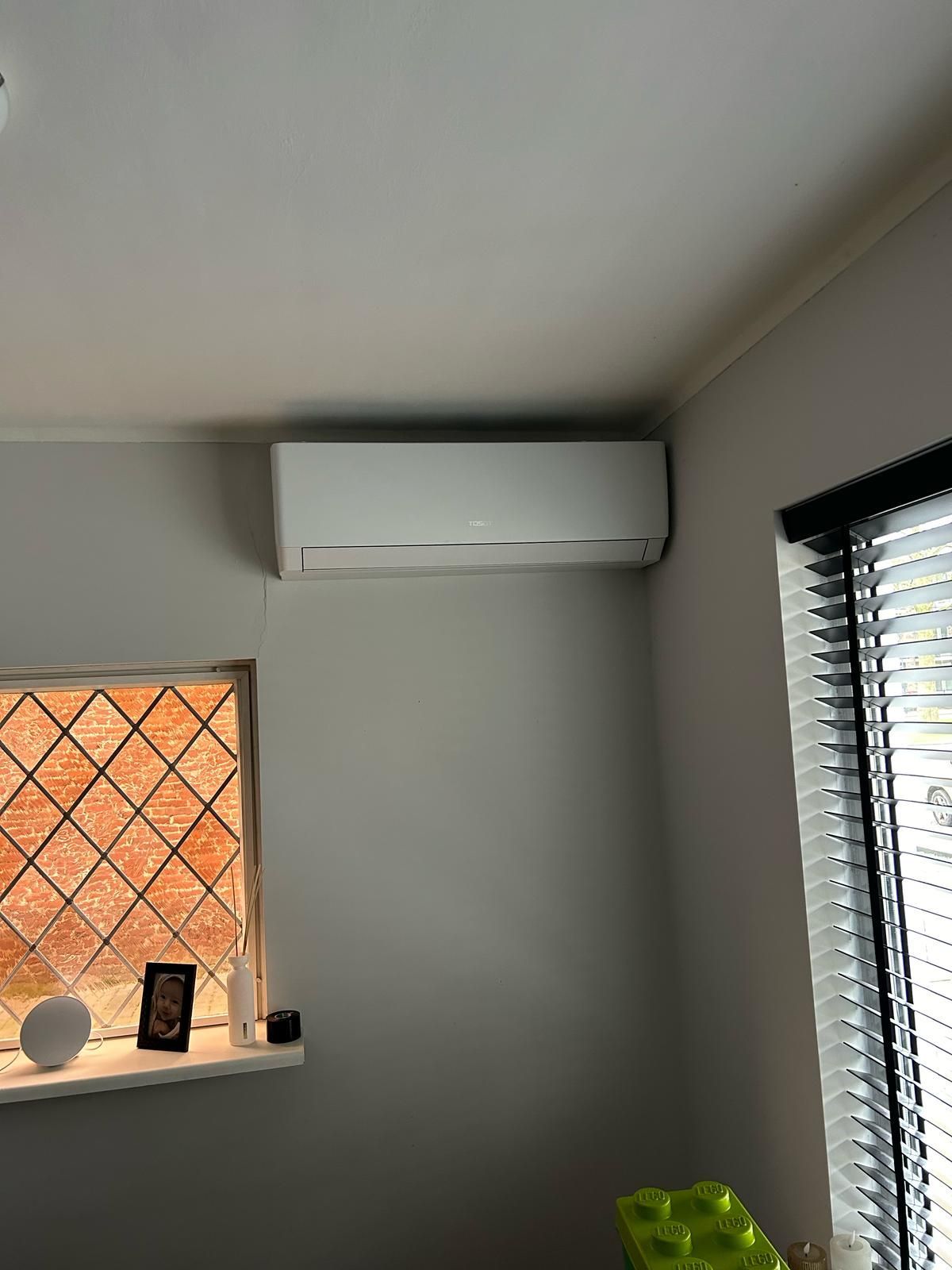 Witte airconditioningunit op een grijze muur boven een raam met jaloezieën.