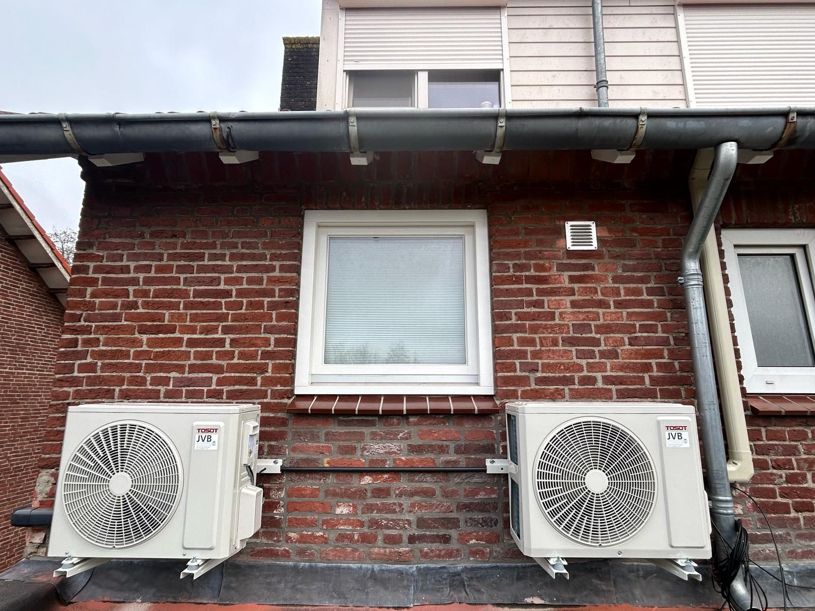 Twee airconditioningunits gemonteerd aan een bakstenen muur onder een raam, met een dakgoot erboven.
