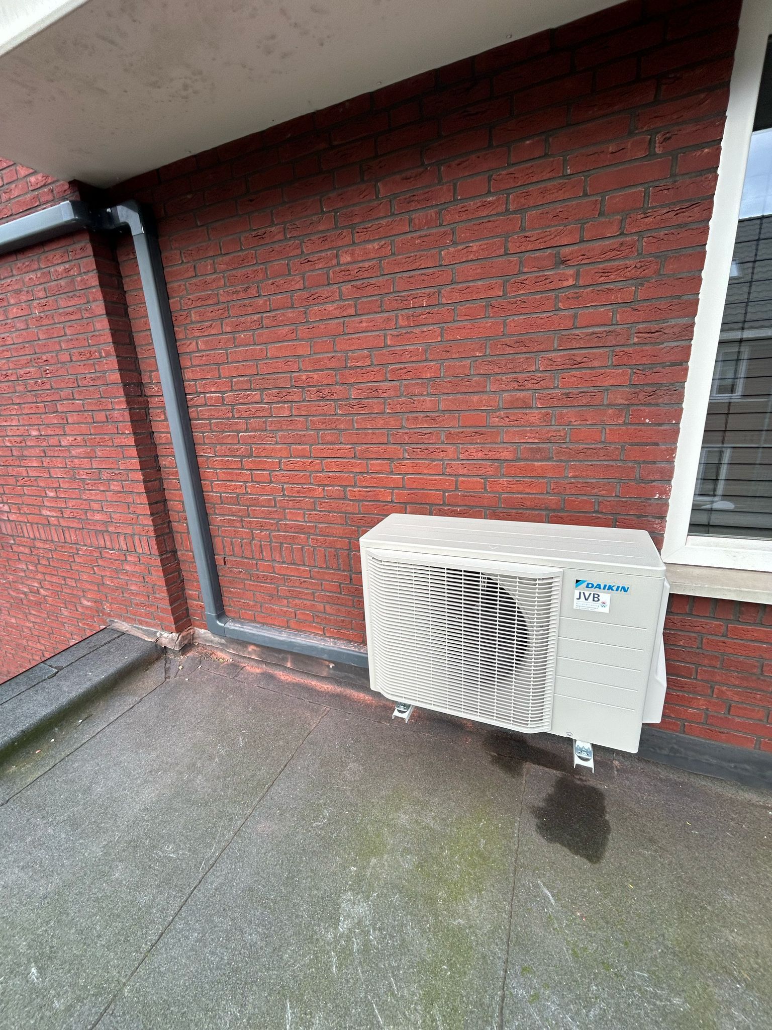 Airconditioningunit gemonteerd op een bakstenen muur naast een raam en een dakgoot.