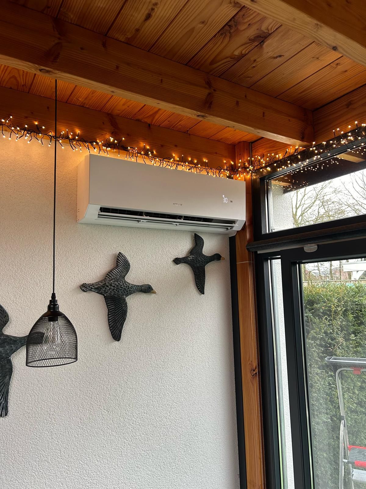 Airconditioning gemonteerd op een witte muur met structuurverf, versierd met vogelkunst en lichtslingers.