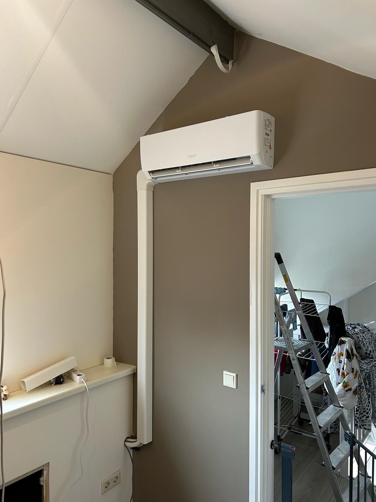 Een witte airconditionerunit gemonteerd aan een bruine muur, met witte leidingen en een deuropening naar een rommelig interieur.