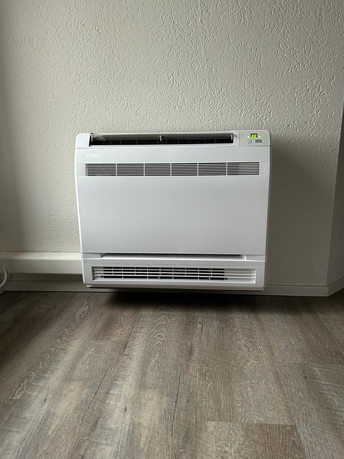 Witte, aan de muur gemonteerde airconditioningunit op een gespikkelde muur boven een vloer met houtlook.