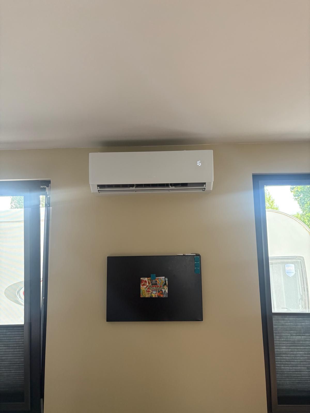 Airconditioning gemonteerd aan een beige muur boven een zwarte rechthoek, geflankeerd door ramen met jaloezieën.