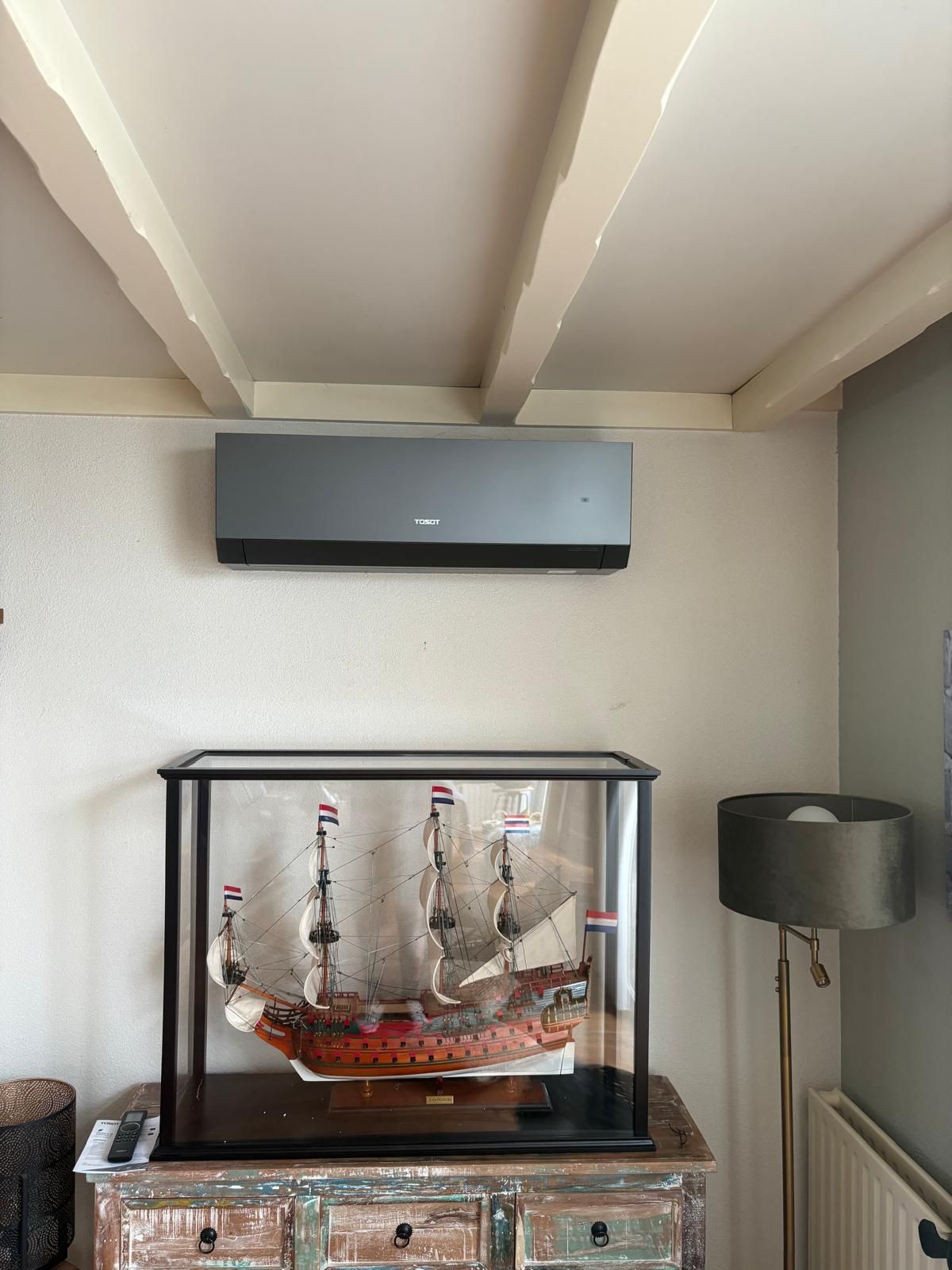 Een airconditioner is boven een vitrine met modelboten gemonteerd in een kamer met witte balken en een lamp.