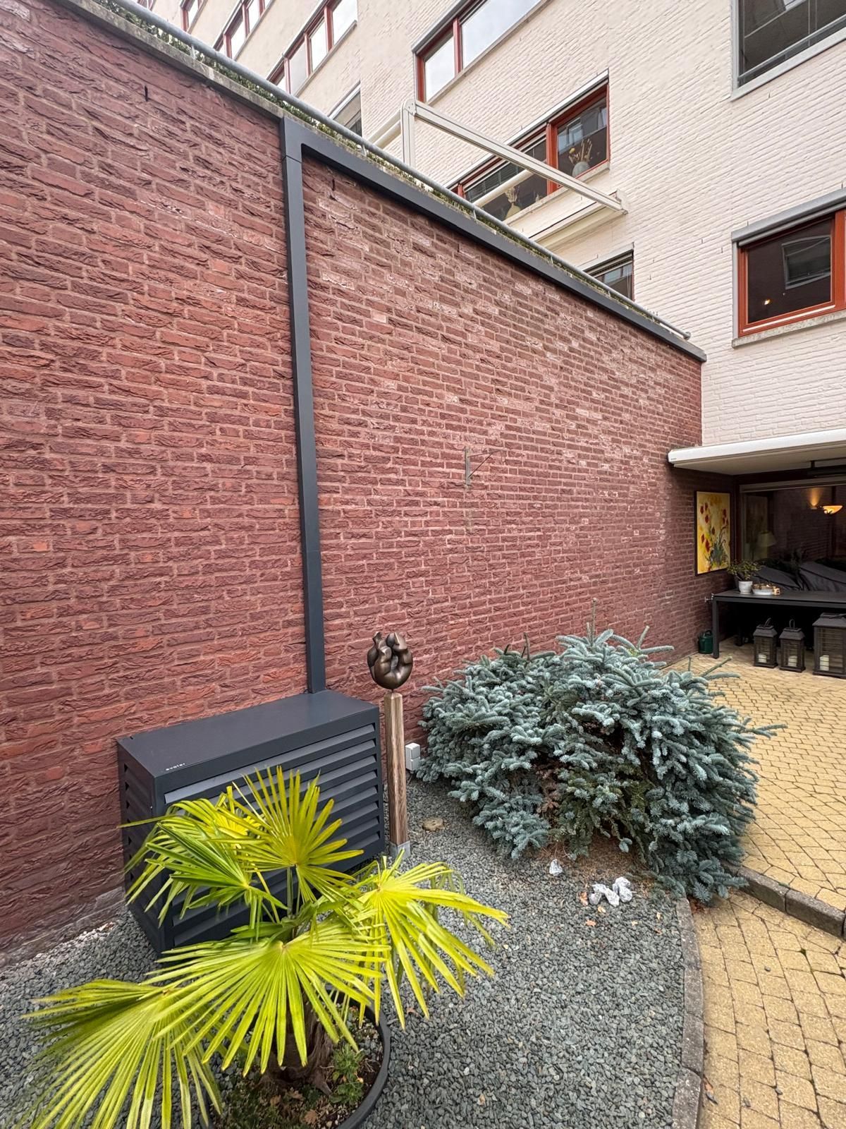 Een rode bakstenen muur met zwarte sierlijsten, een kleine tuin met een blauwe struik en een palmboom.